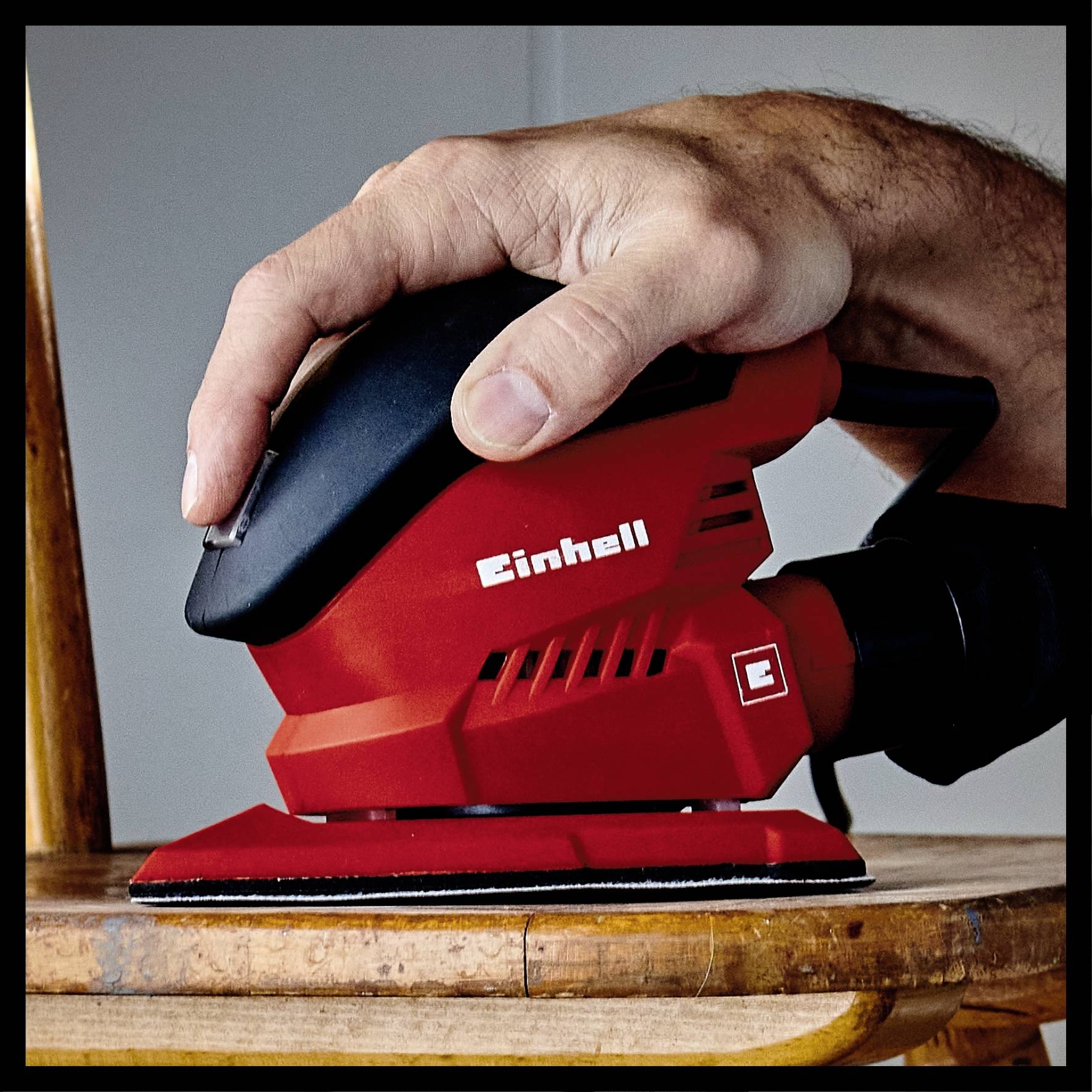A person is sanding a wooden chair with a red orbital sander. The hand is guiding the tool across the surface of the chair.