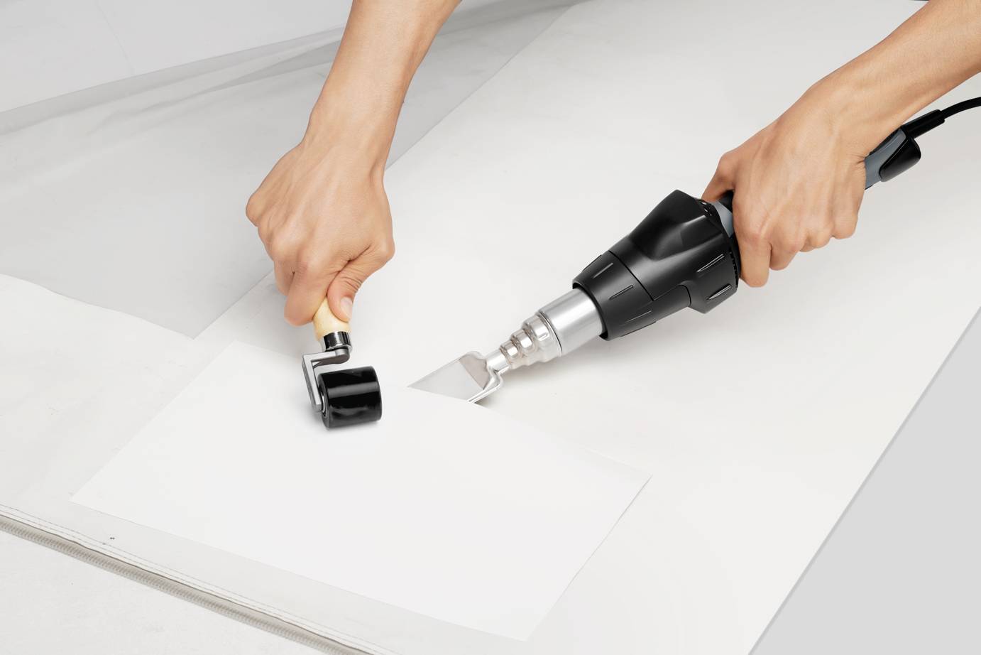 Two hands using a hot air gun and a roller to weld plastic material.