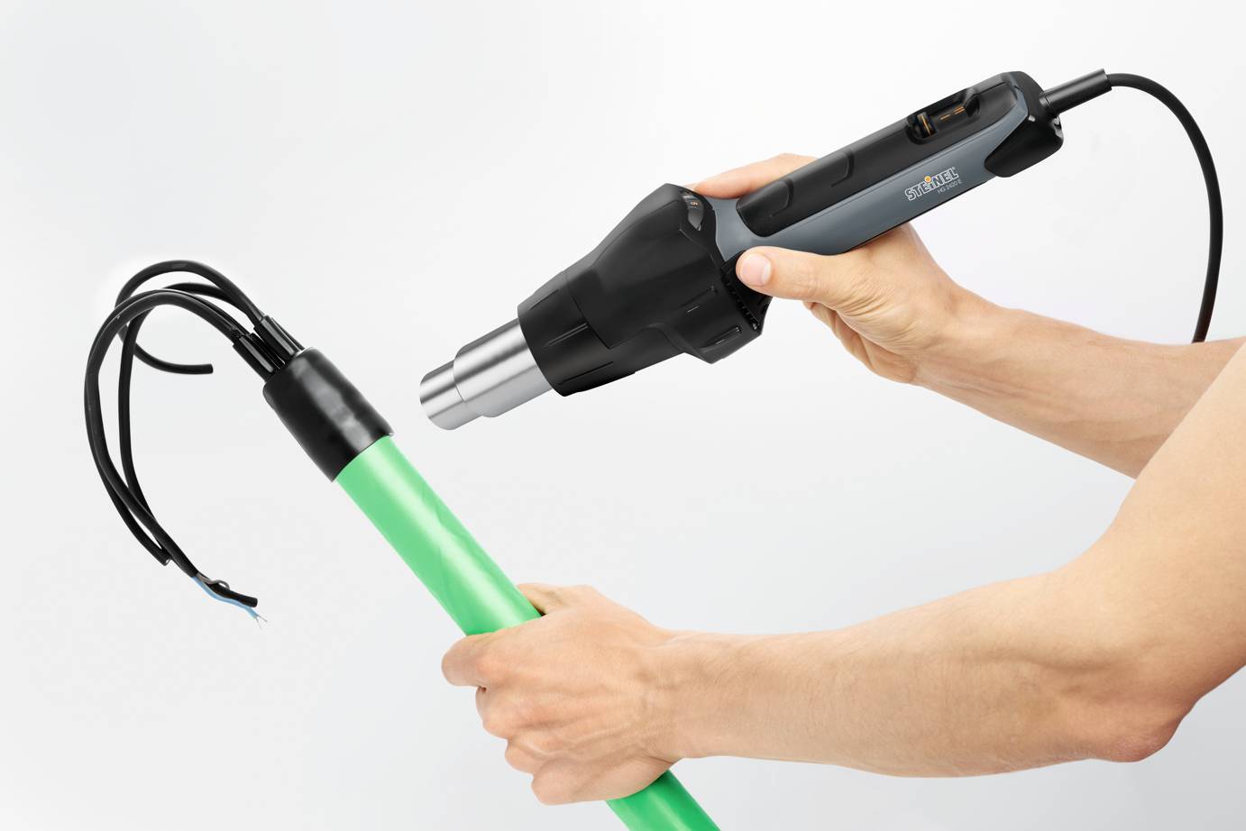 A person is holding a hot air blower and using it to heat the end of a green pipe, with black cables protruding from it.
