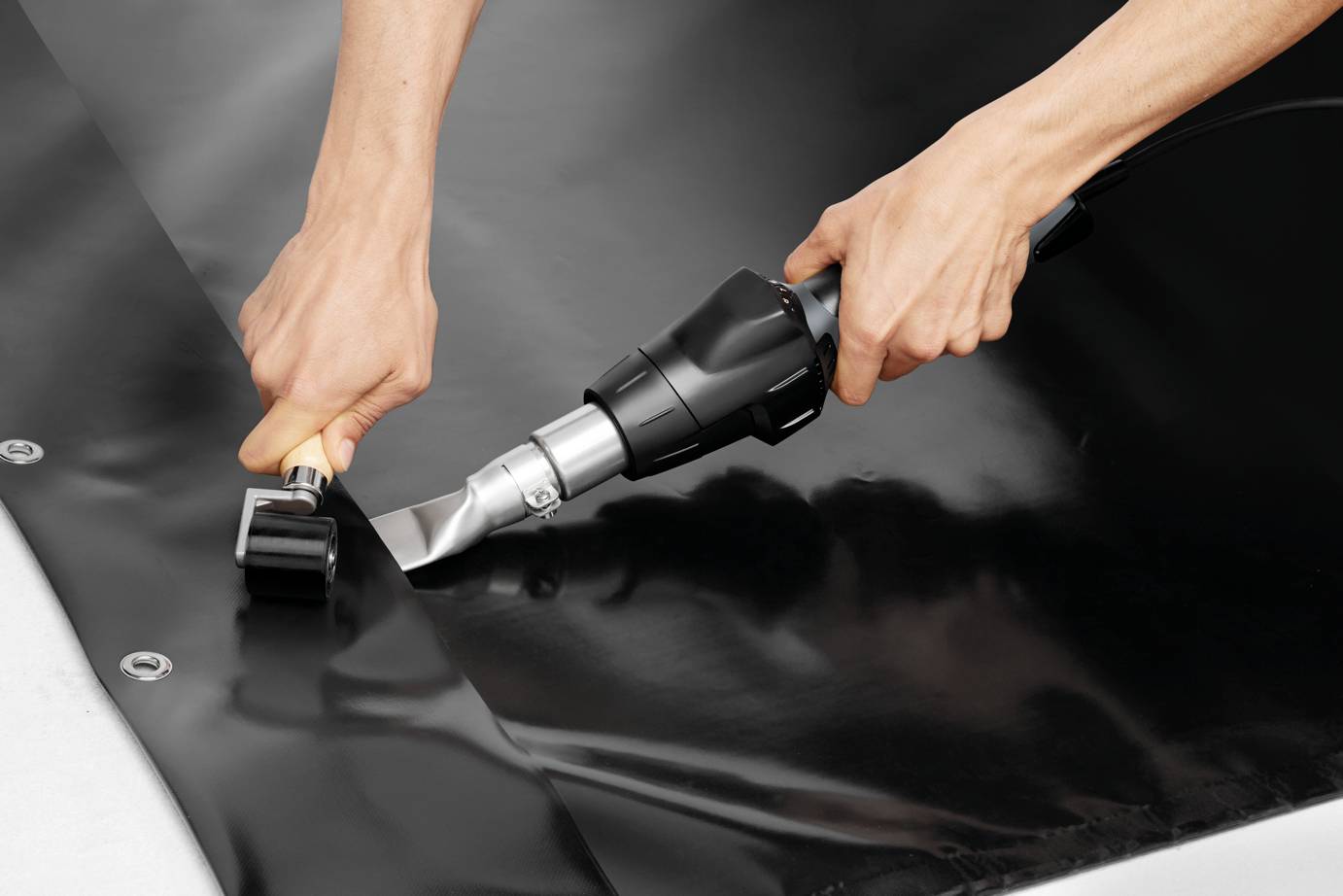 Two hands are using a hot air welding device to weld black plastic film onto a metal table.