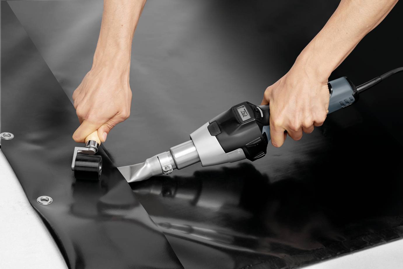 'A person is welding two black tarpaulin sections with a handheld welding device. Their hands are holding the device securely and guiding the weld seam.'