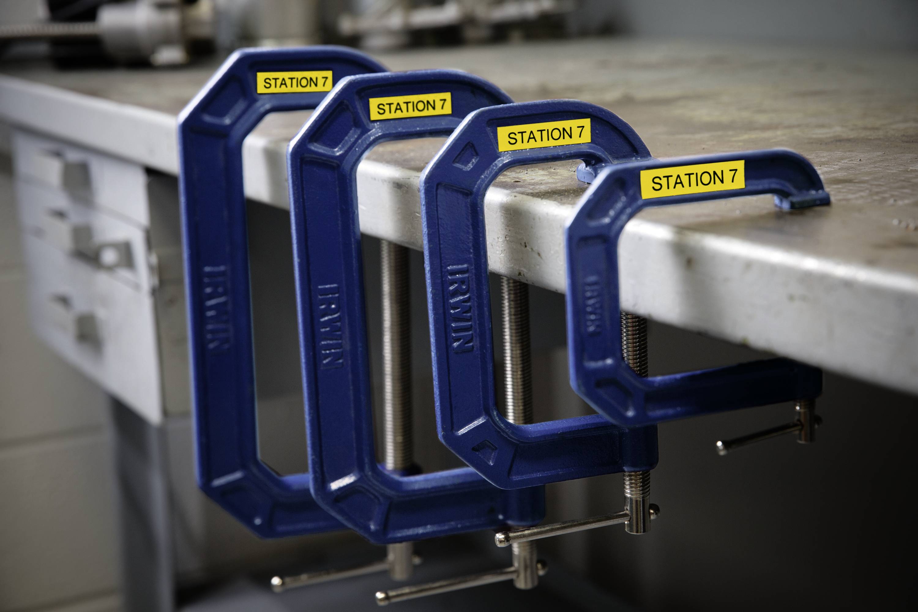 Four blue G-clamps of different sizes are clamped to a workbench edge. Each clamp is labelled 'Station 7'.