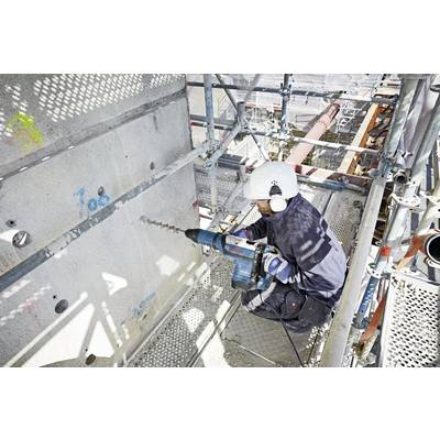 A worker in safety gear uses a power drill on a concrete wall at a construction site, surrounded by scaffolding, highlighting safety practices.