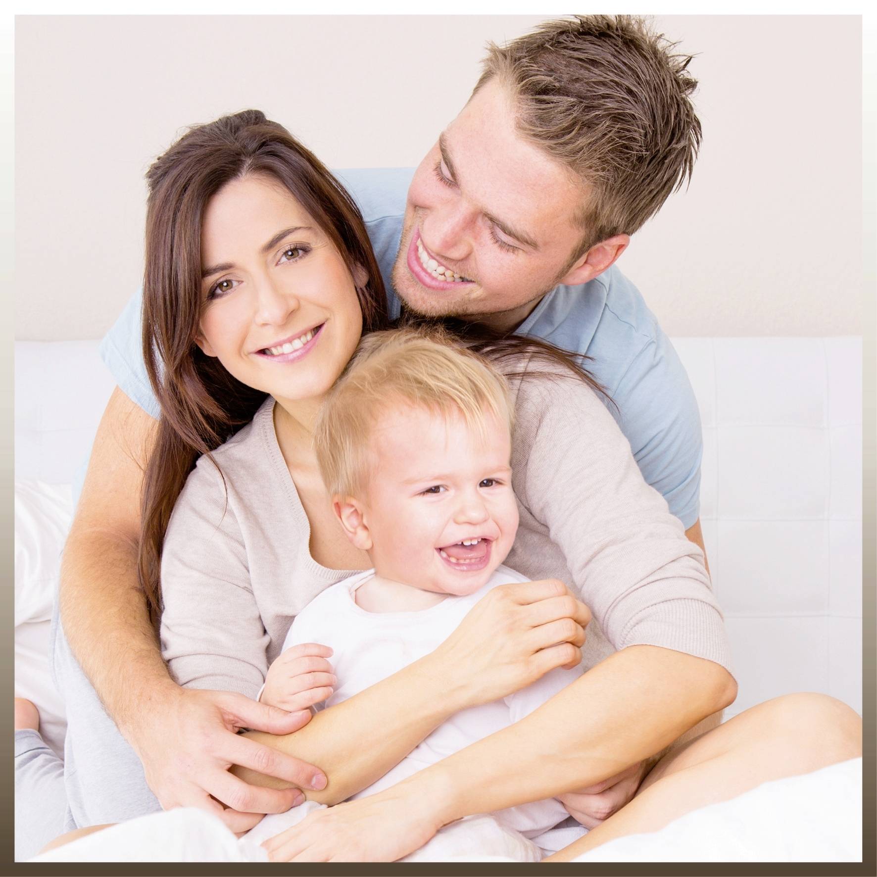 A laughing family sits together on a sofa. A man and a woman are holding a smiling little child in their arms.