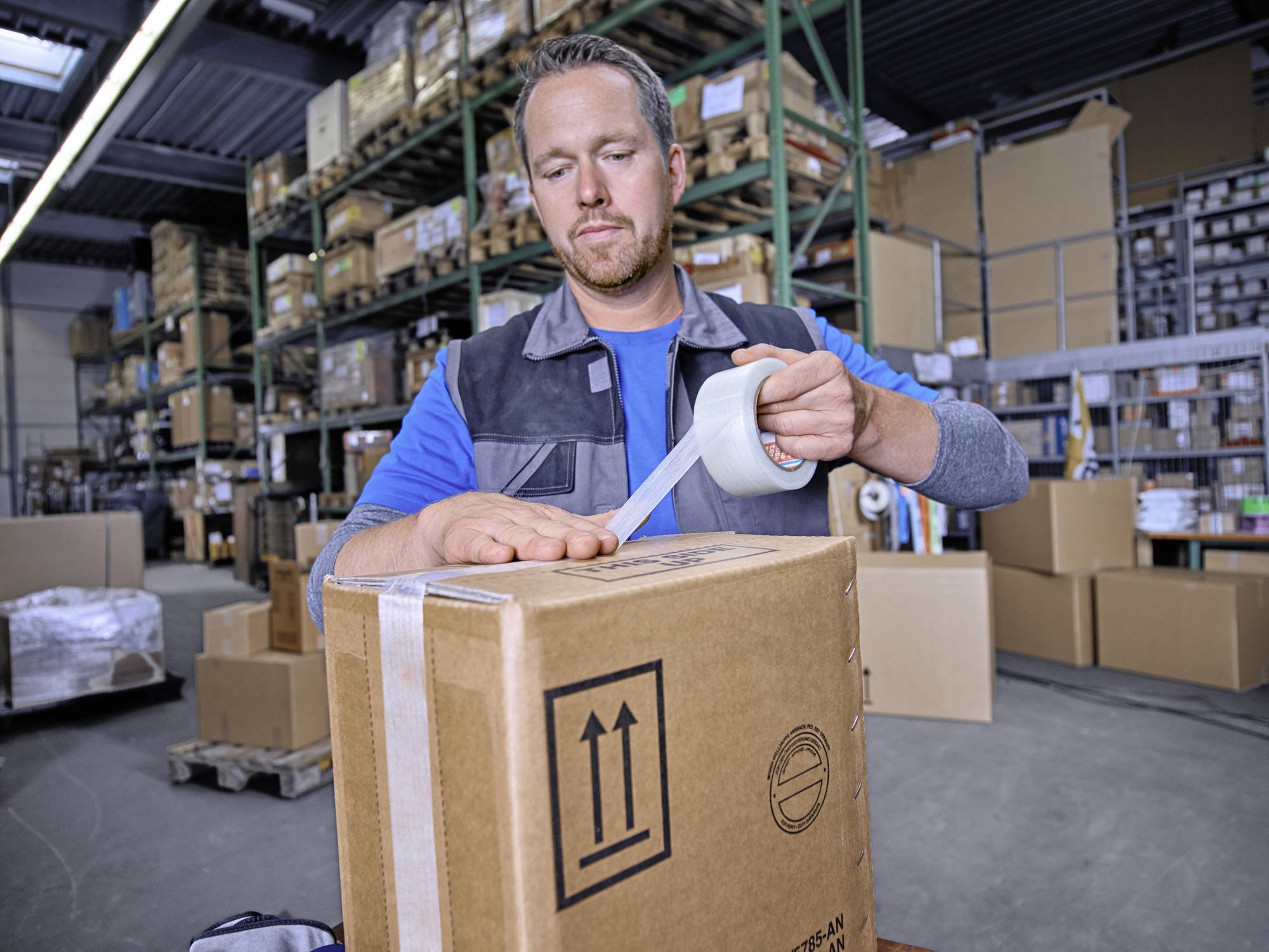 A man is sealing a cardboard box with parcel tape in a warehouse. Storage shelves with additional packages are visible in the background.