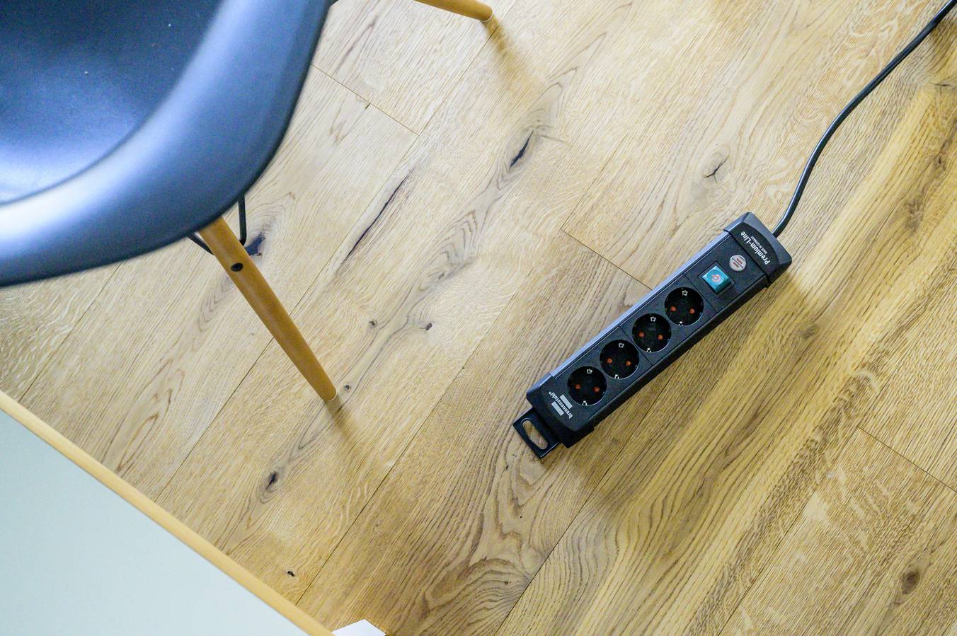 A black power strip with four sockets is lying on a wooden floor next to a chair.