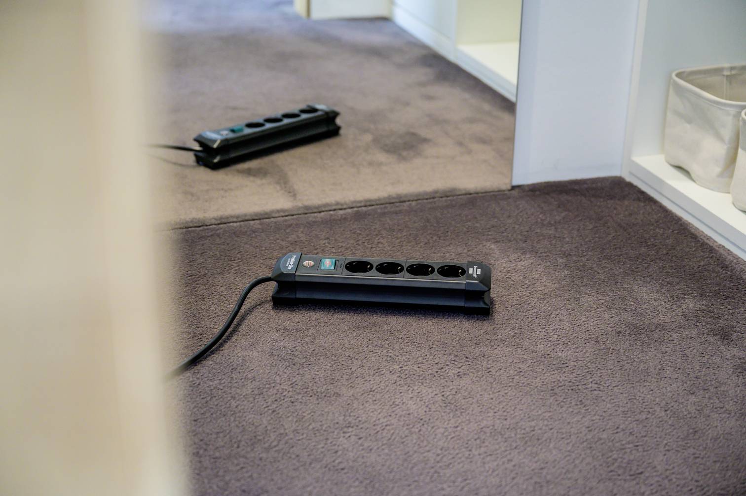 An extension lead with multiple sockets is lying on a carpet, partially underneath an open cupboard.