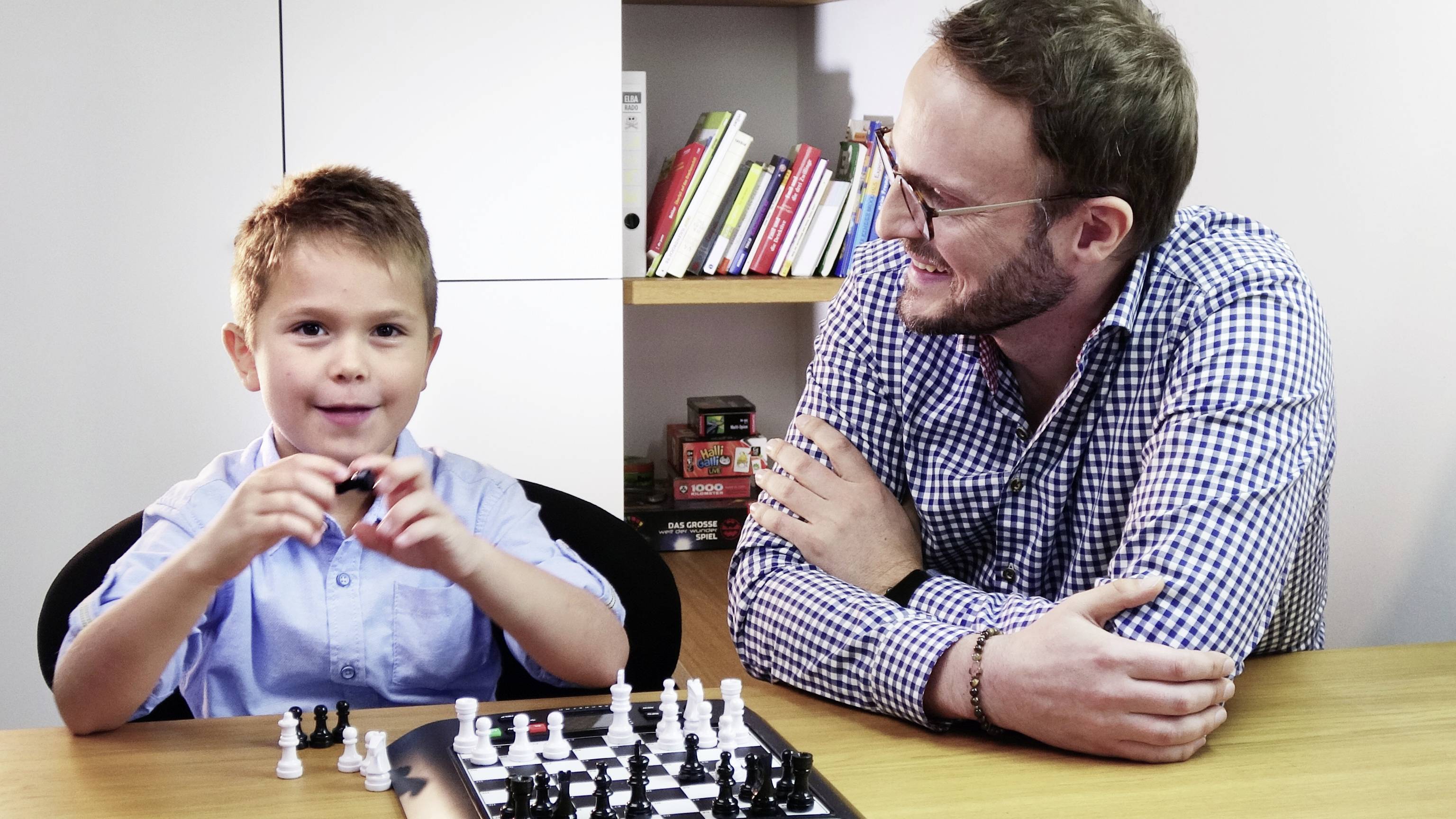 A man and a boy are sitting at a table playing chess. The boy is holding a chess piece in his hand and smiling, while the man looks on kindly.