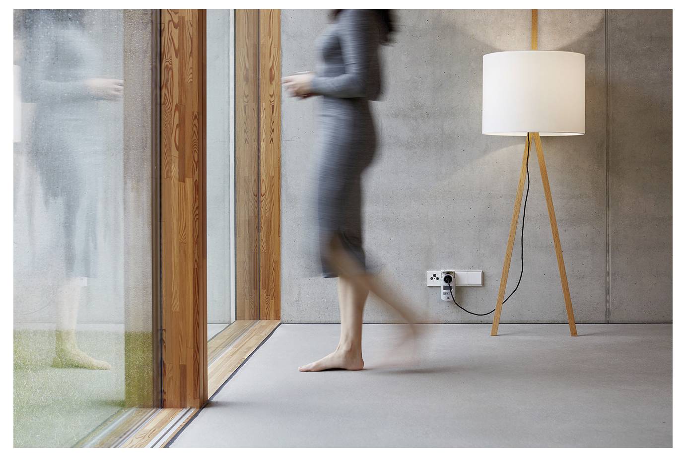 A blurred barefoot person walks past a modern floor lamp in a minimalist room with large glass doors and wood trim.<br>