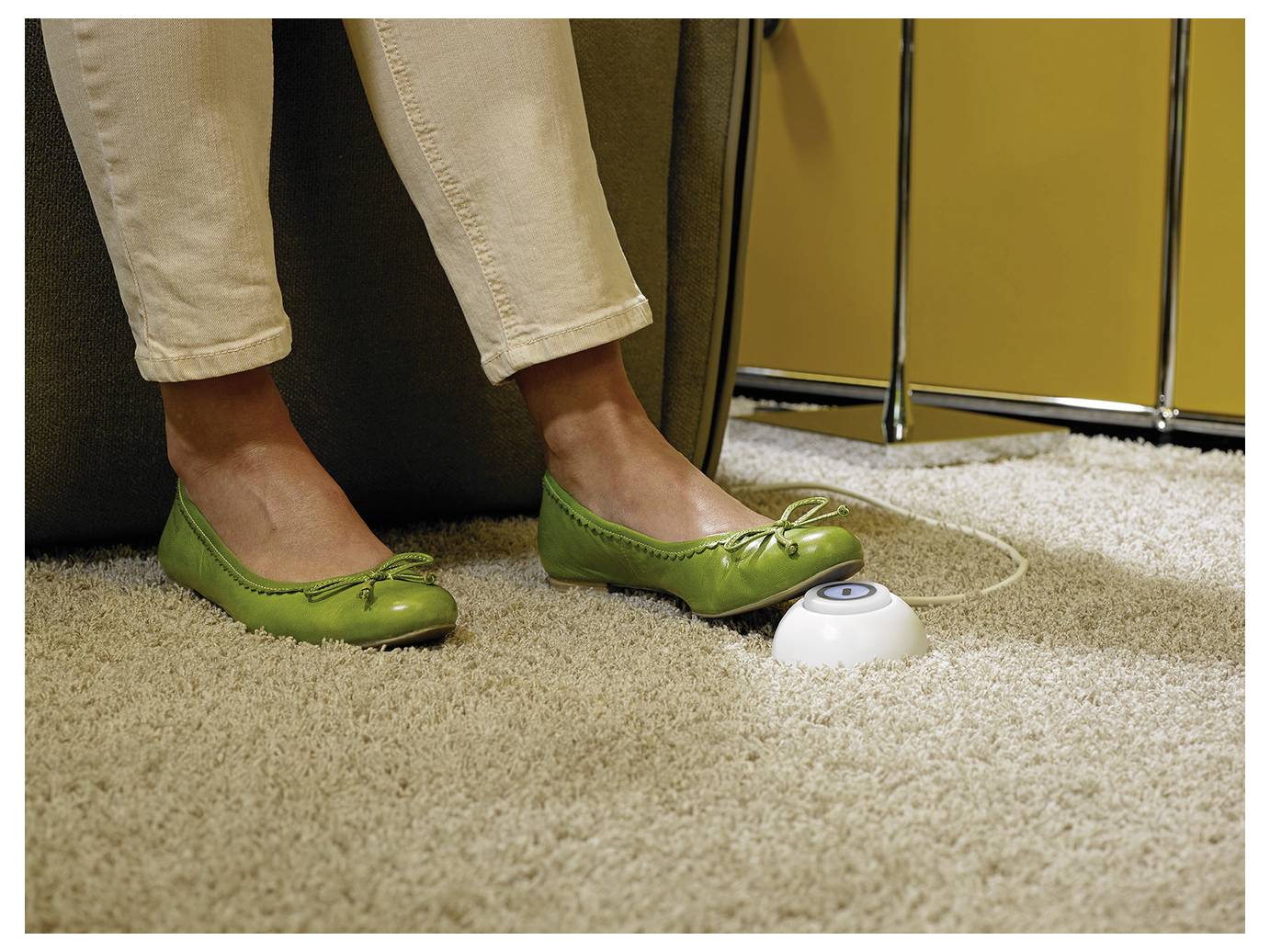 Close-up of feet in green flats resting on carpet near a small white foot pedal or button beside a chair.