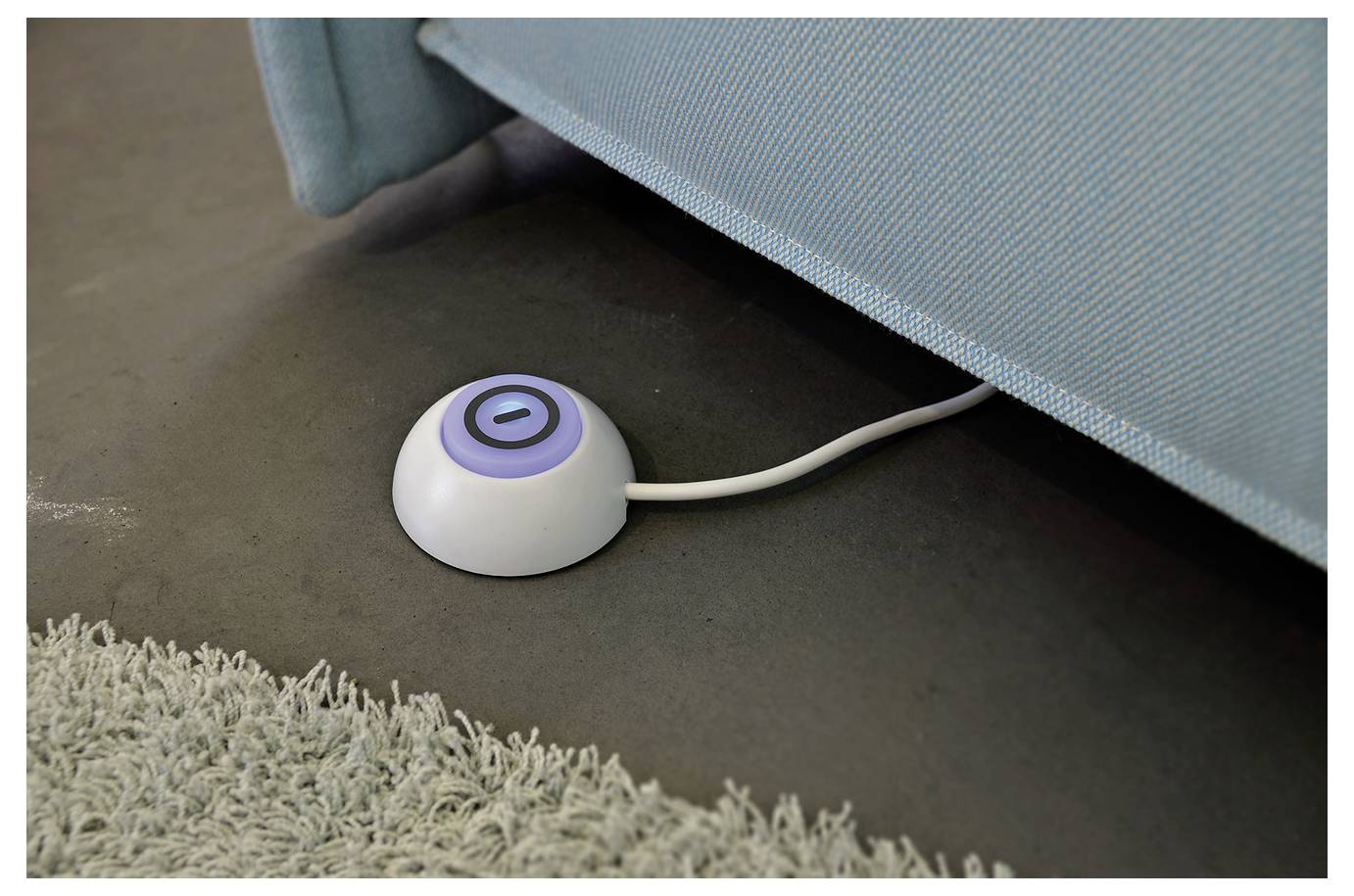 Small round white floor button with a blue power symbol beside a sofa, likely a hidden control or floor switch.