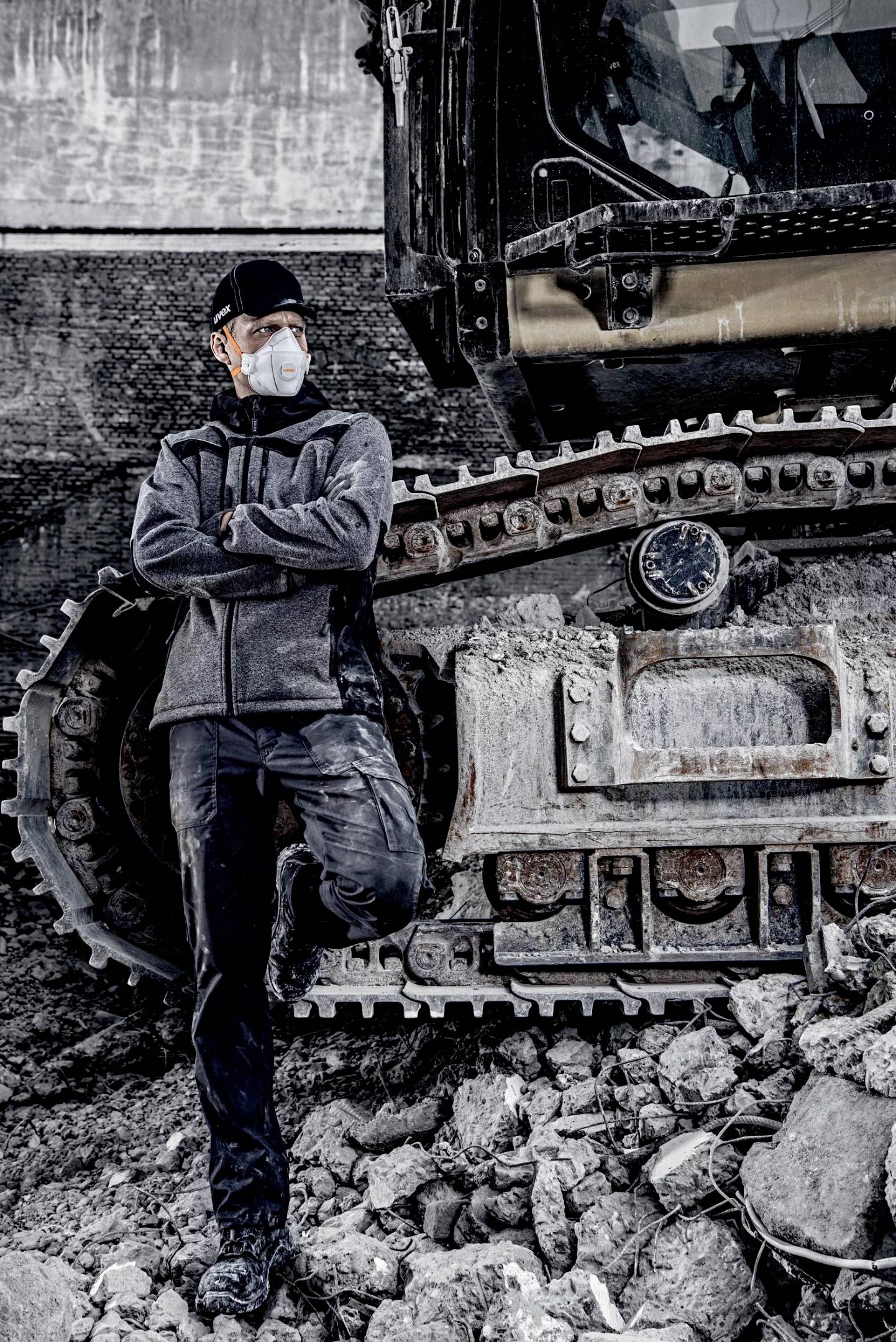 A worker wearing a face mask leans against a large excavator on a construction site. He is dressed in dark workwear and covered in dust.
