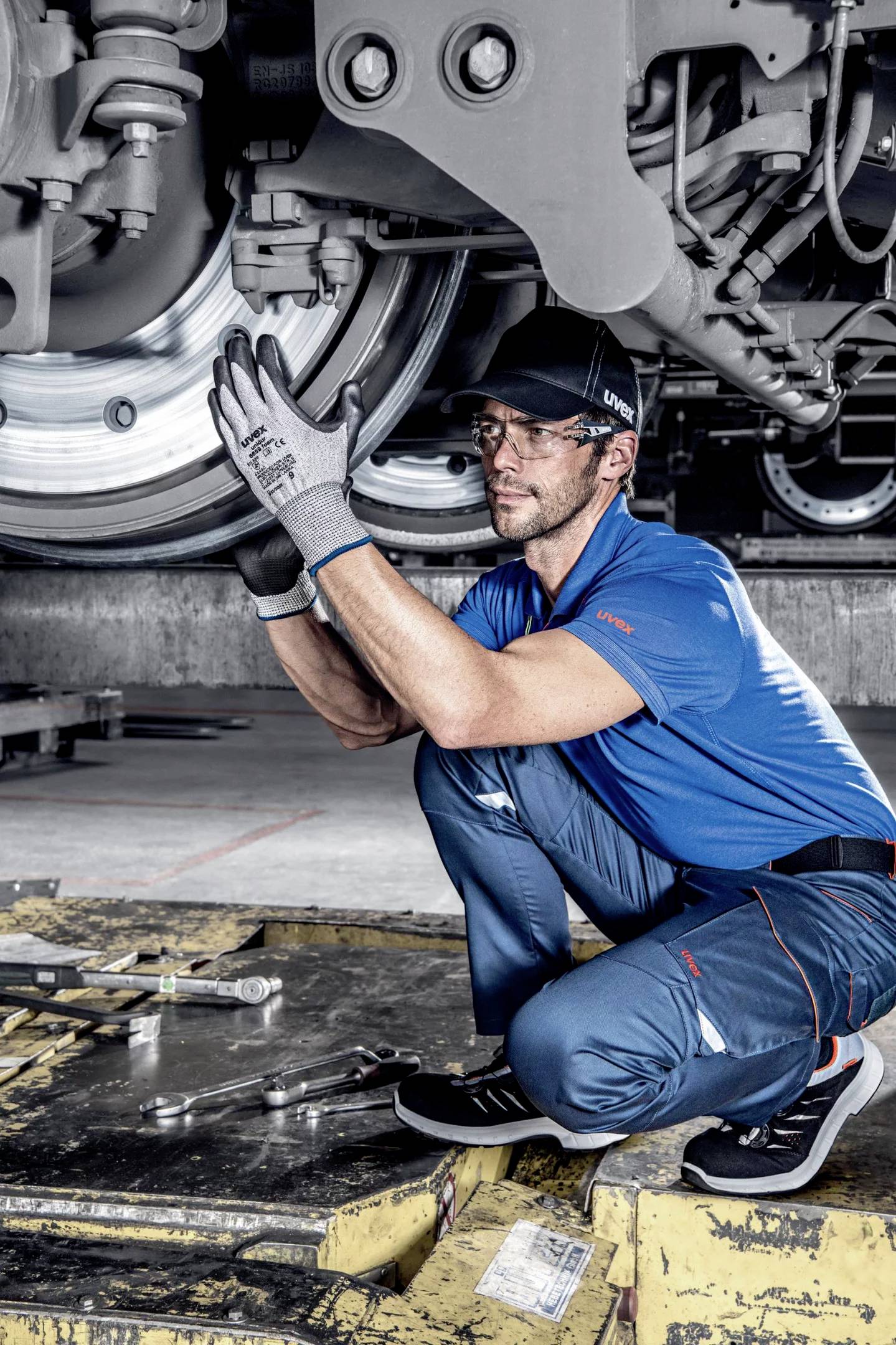 A mechanic in blue overalls is inspecting the undercarriage of a train. He is wearing gloves and carefully examining the mechanical components.