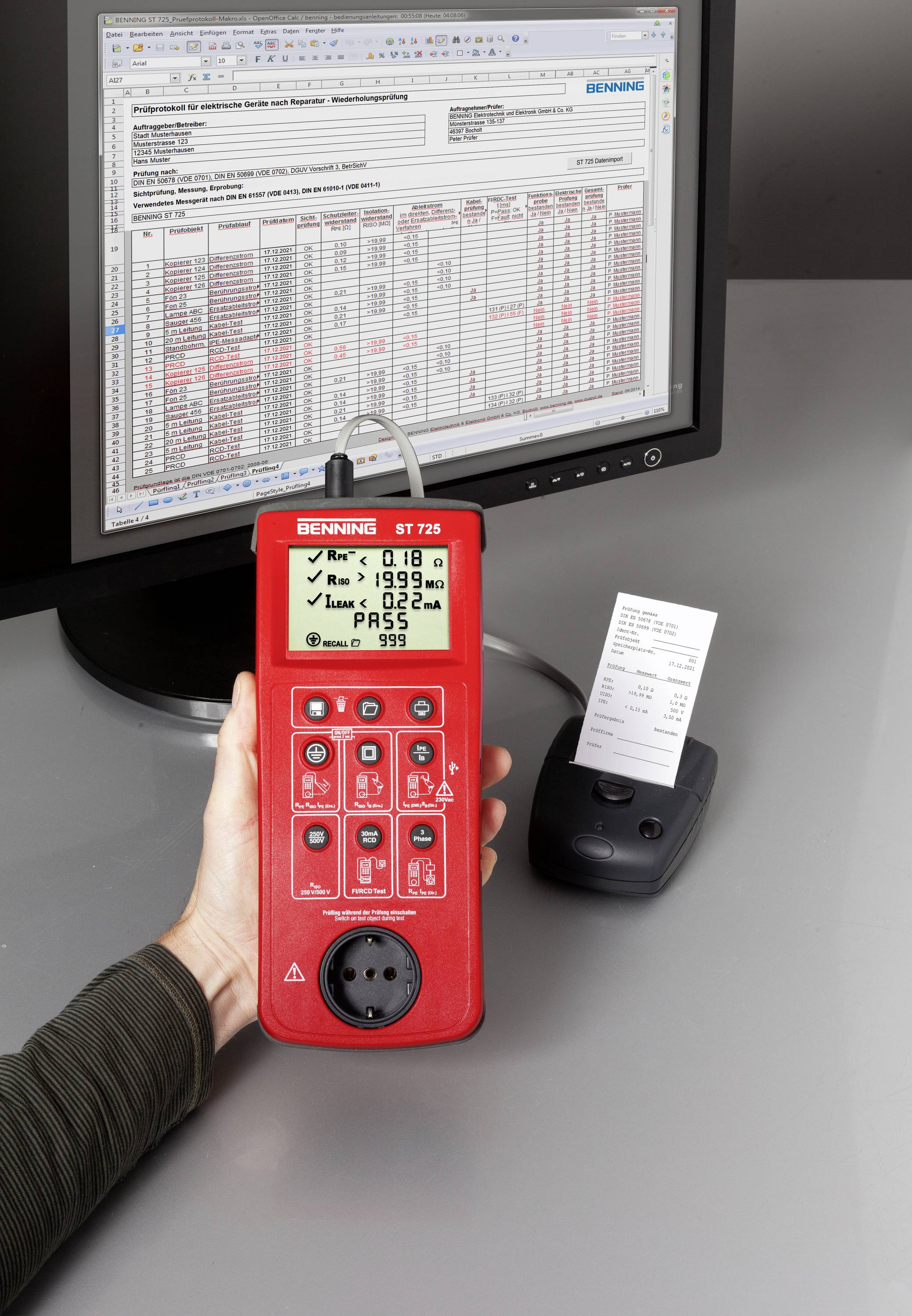 A hand holds a red testing device in front of a computer screen displaying a table of measurement results. A printout lies beside it.