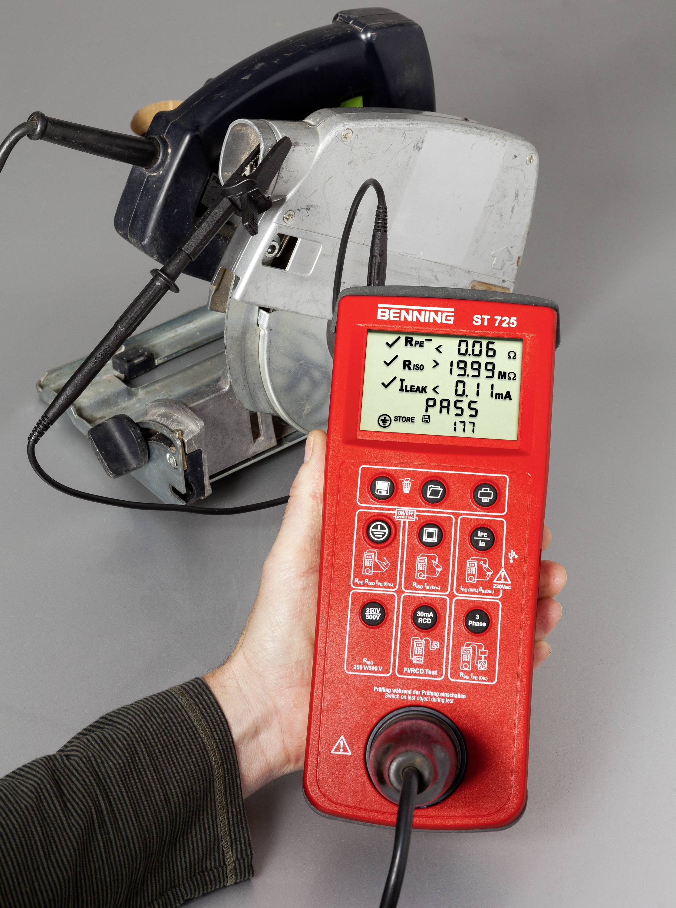 A person is holding a red electrical testing device connected to a damaged power tool to test its safety.