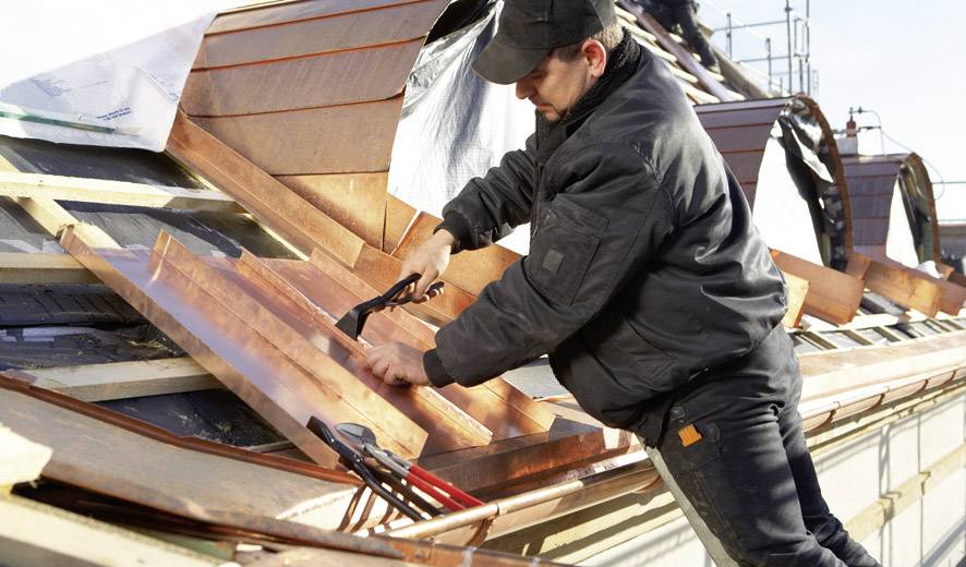 A worker in protective clothing is securing copper plates on a roof. Scaffolding is visible in the background.