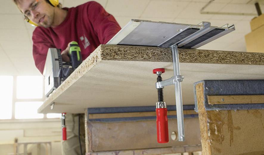 A craftsman is sawing a chipboard with a circular saw in a workshop. The board is secured with clamps.