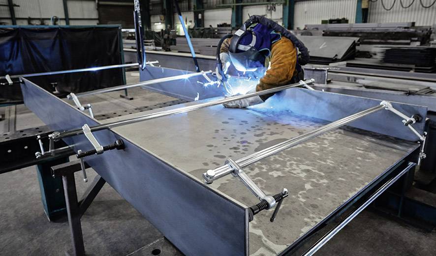 A welder in protective clothing is working on a metal piece in a workshop, surrounded by clamps and equipment, with light flashes visible.