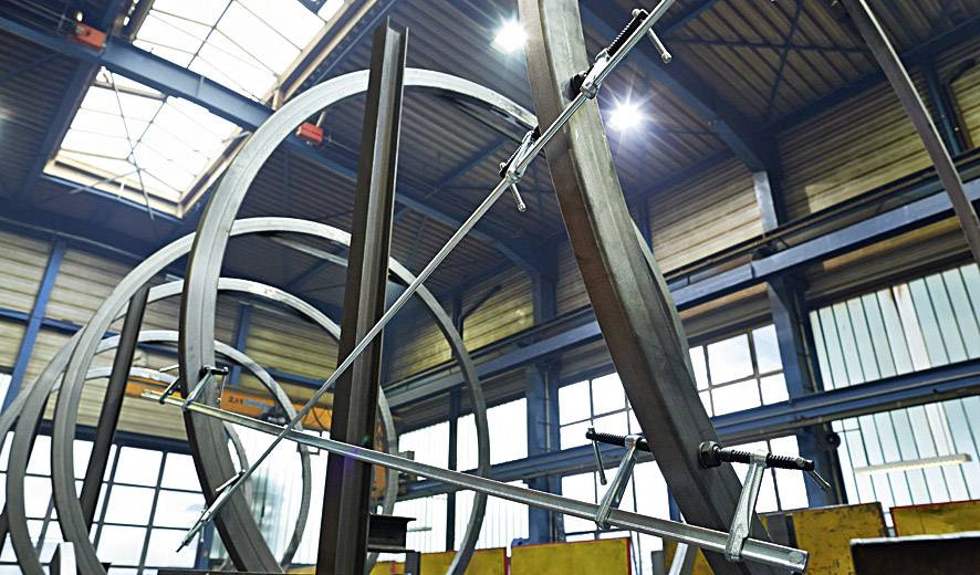 Steel sculpture under construction in a workshop hall with visible roof structure and natural light; metal beams in rounded forms.