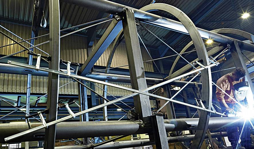 A person is welding a large steel frame in a factory hall. Metal structures and tools are visible in the background.