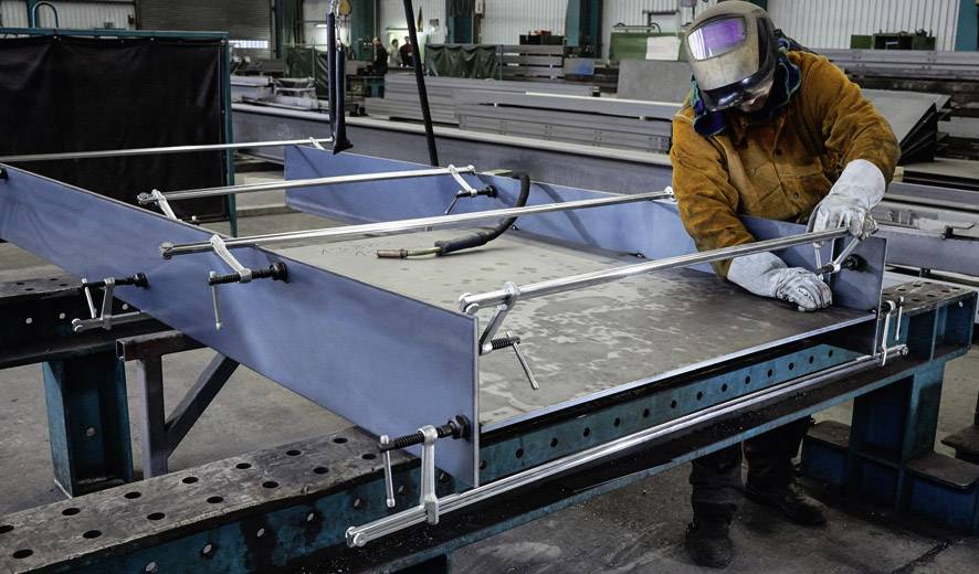 A person wearing protective clothing and a welding helmet is working on a metal construction in a factory hall.