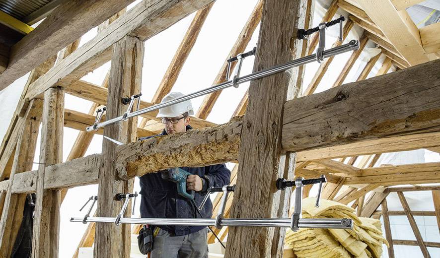 A construction worker wearing a hard hat is working with a tool on a wooden roof structure. Several props are supporting the beams.