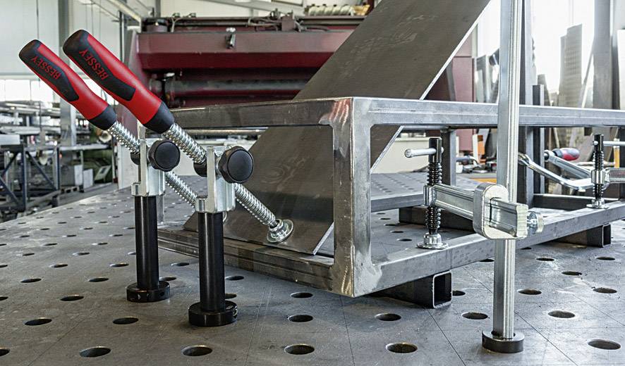 Metal workshop with a fixed metal frame mounted on a perforated metal table. Several clamps hold the frame in place during processing.