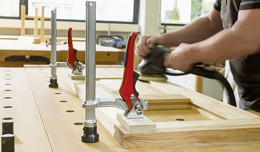 A person is sanding a wooden frame on a workbench, secured with two red G-clamps.