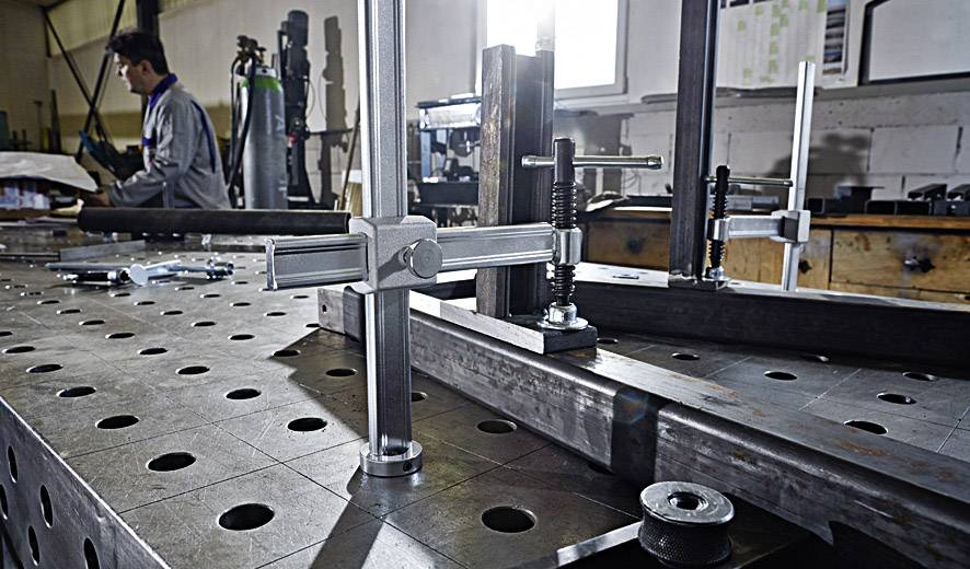 A workshop with a welding table in the foreground; a man is working in the background.