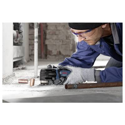 Construction worker using a Bosch hand power tool to cut a copper pipe on a concrete floor, wearing gloves and safety glasses.
