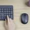 A person is using a wireless mouse next to a keyboard on a desk. Beside it is a bowl of green snacks.