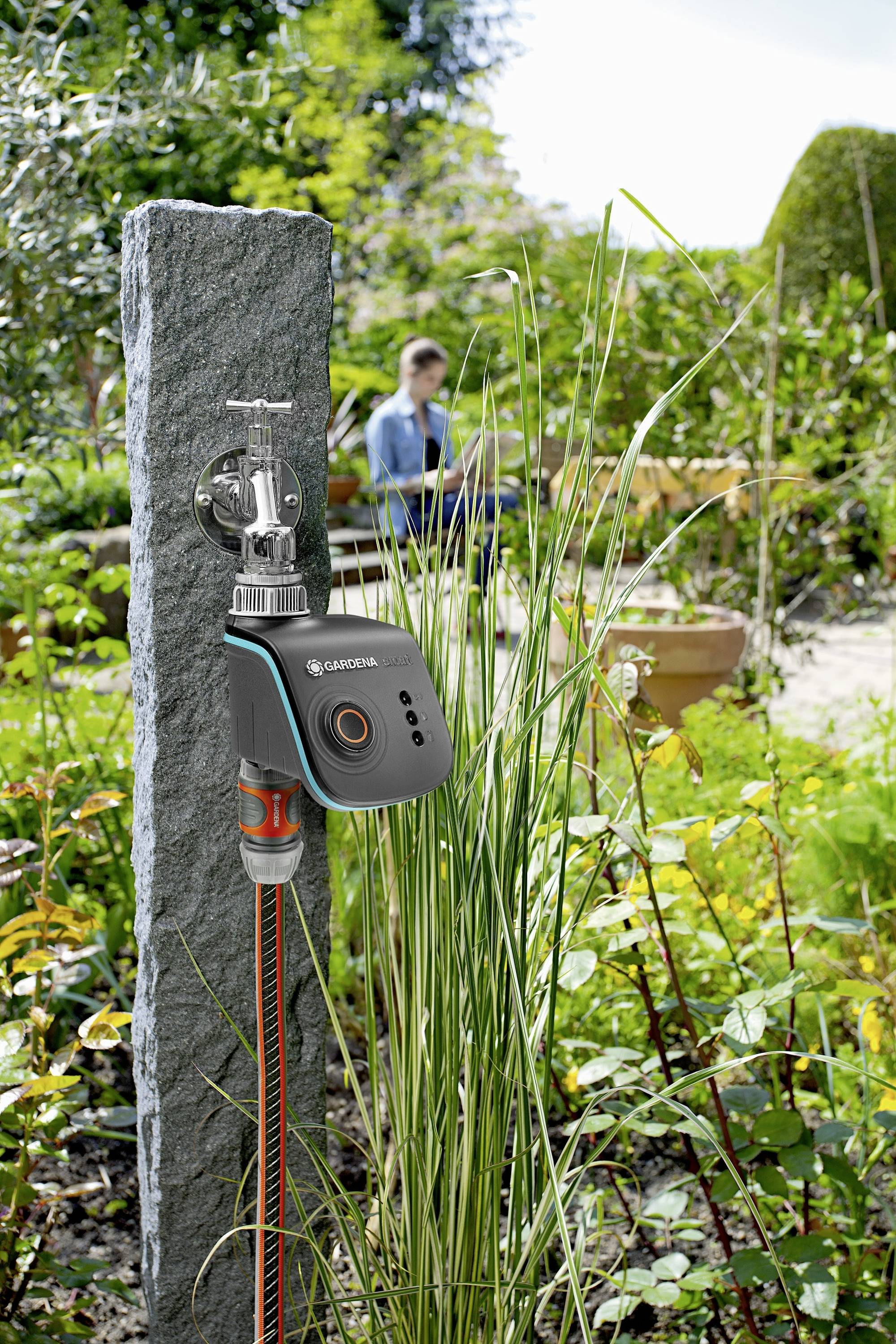 A Gardena irrigation timer is attached to a garden tap. In the background, a person is relaxing in the garden.