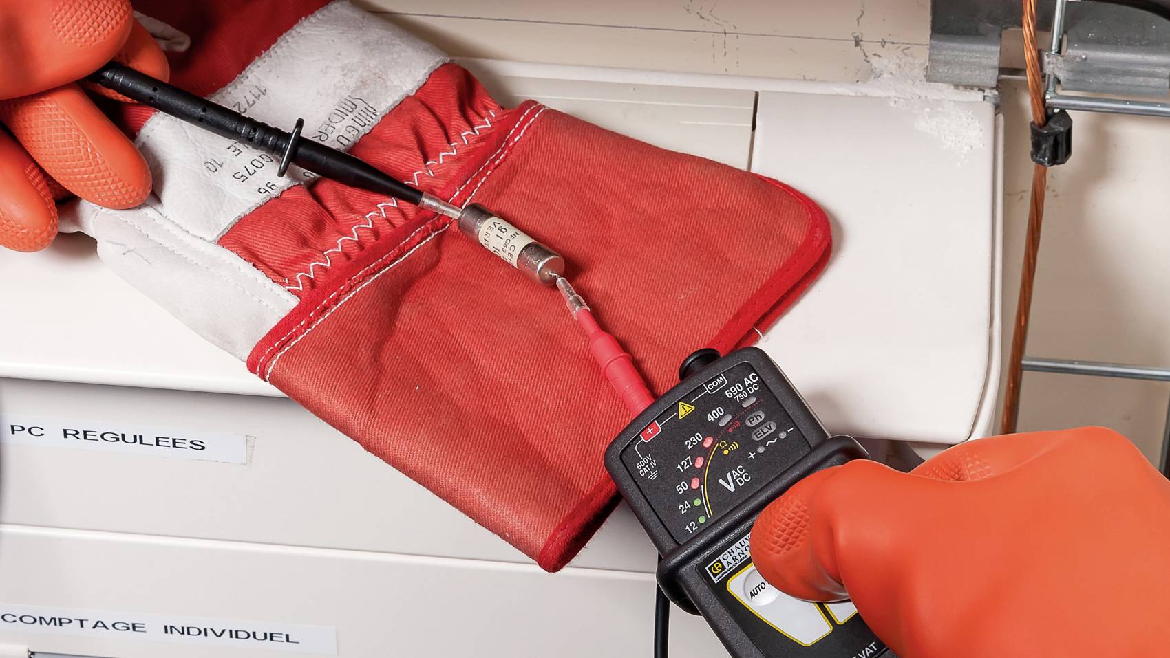 A person wearing orange gloves is measuring the voltage of a cable wrapped in a red protective band using a multimeter.