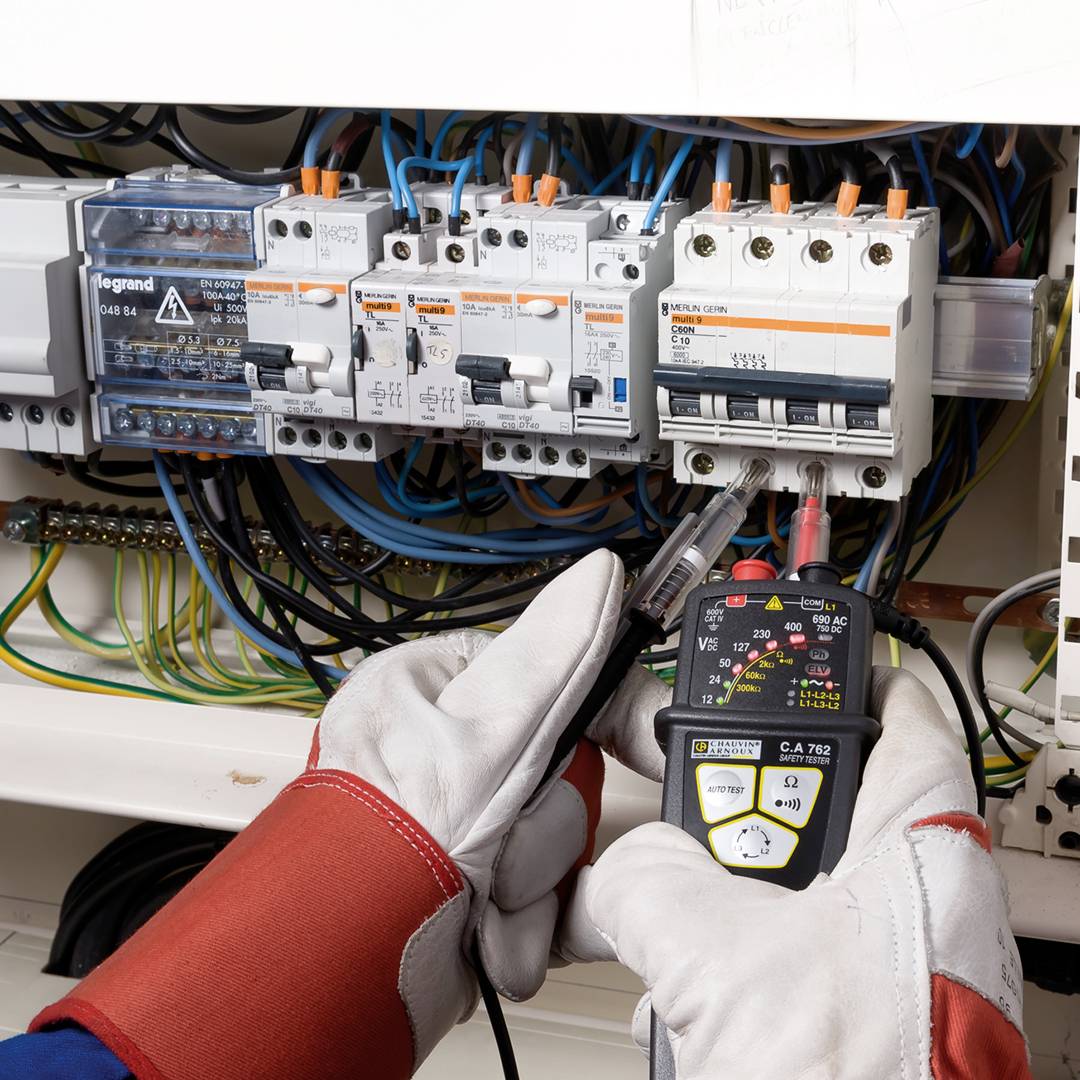 An electrician wearing red gloves checks a fuse in the electrical cabinet using a multimeter.