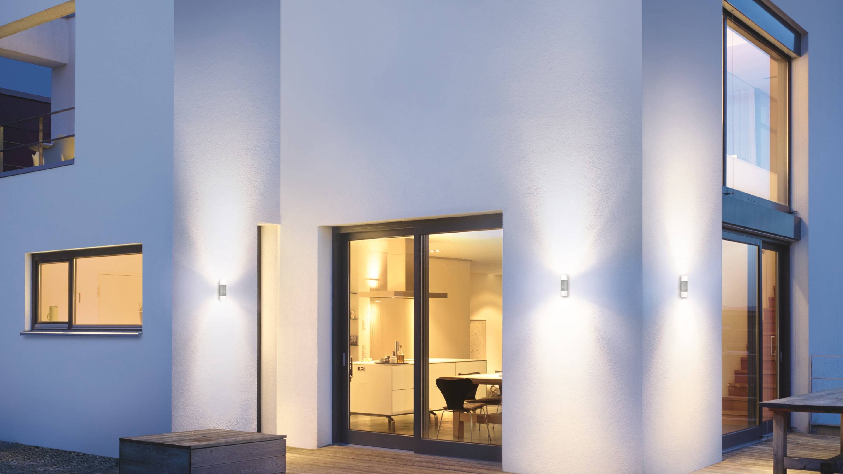 A modern, well-lit house with large windows and a dining area visible from the outside, captured at dusk.