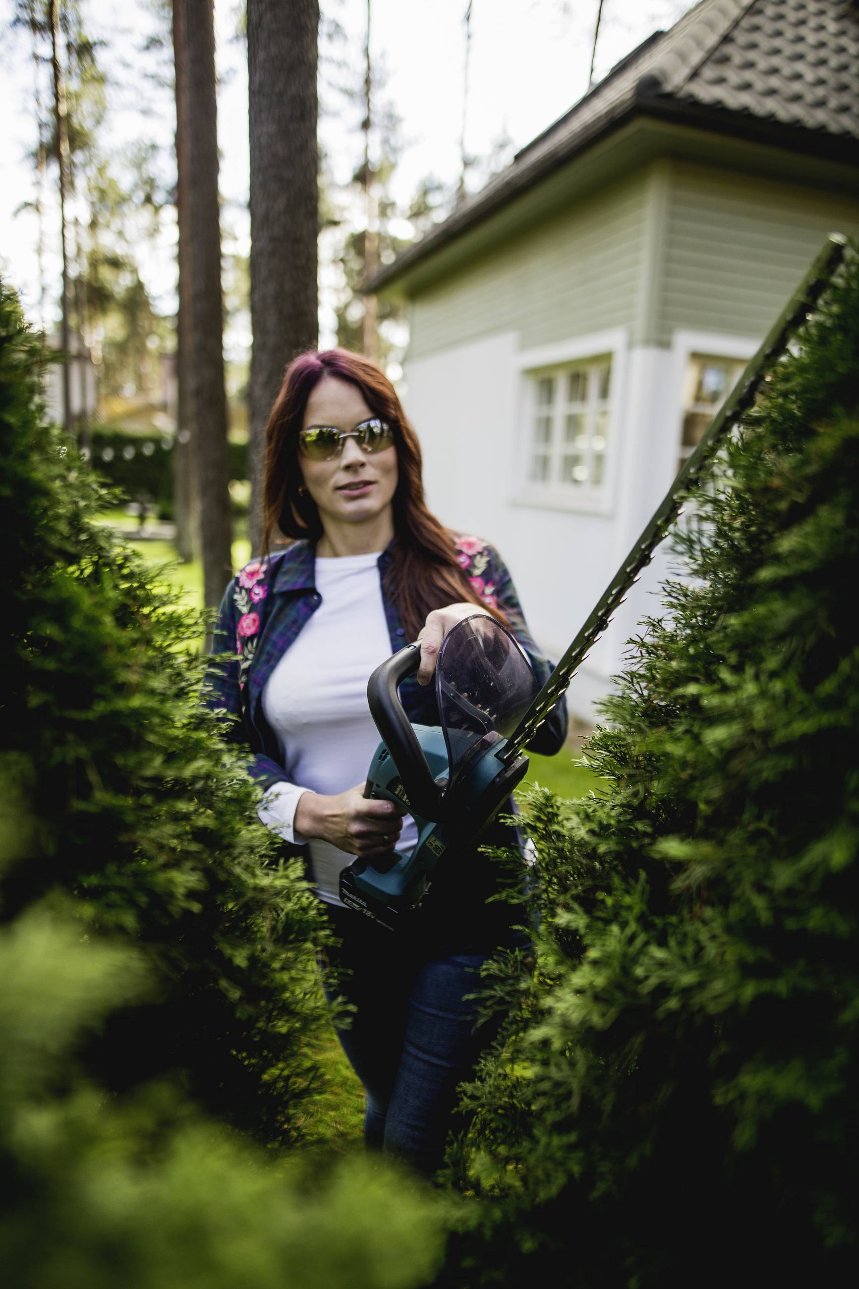 A woman wearing sunglasses is trimming a hedge with an electric hedge trimmer in a garden. A house is visible in the background.