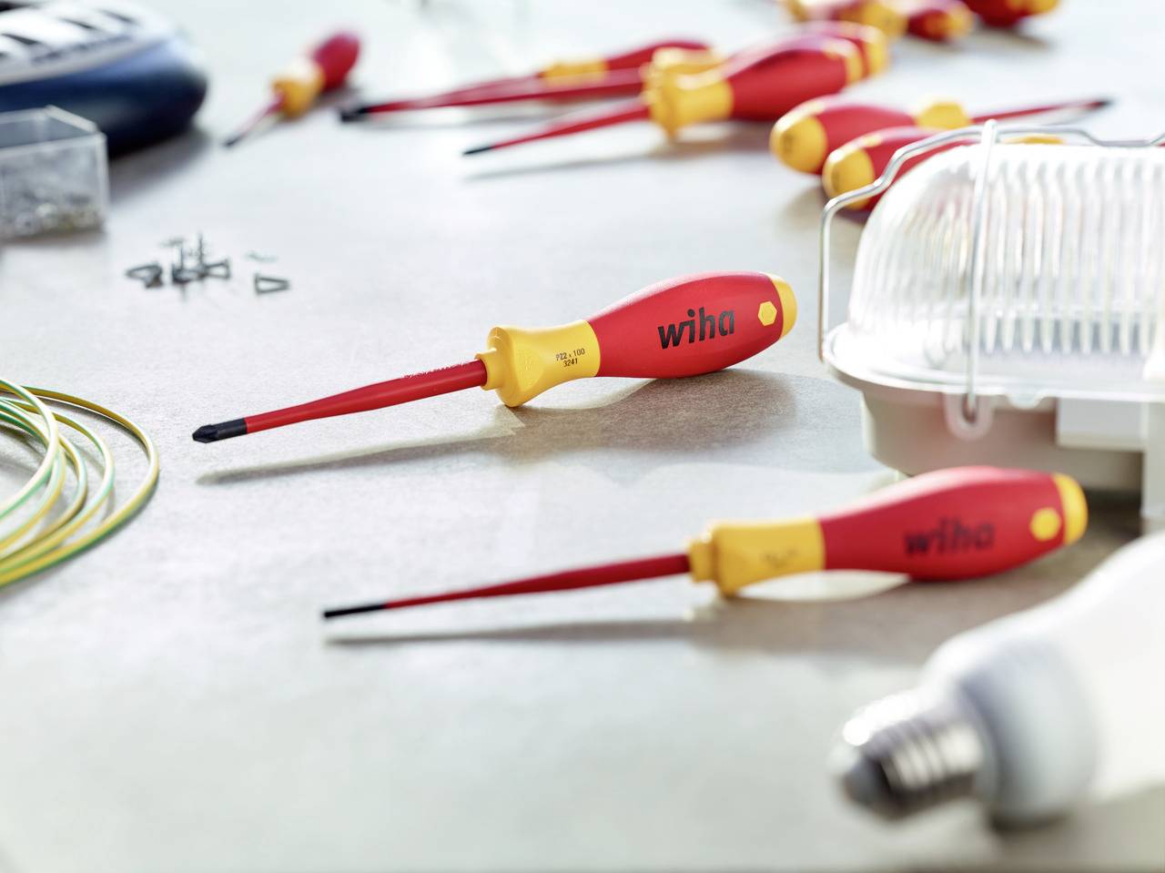 A table with various screwdrivers, cables, and small components. In the foreground, a red screwdriver with a yellow handle lies on the surface.
