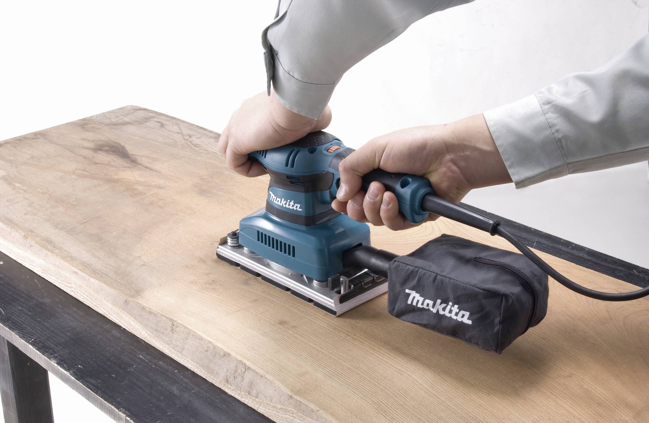 A person is sanding a wooden tabletop with an electric sanding machine. The device is blue and equipped with a dust bag.