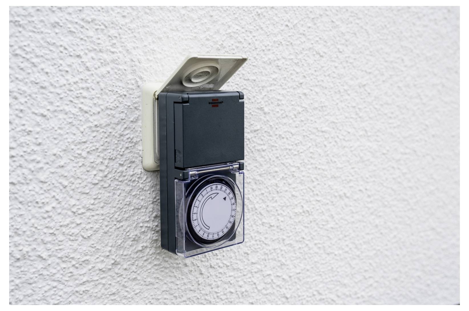A wall-mounted electrical timer switch with a dial and clear cover on a textured white wall.