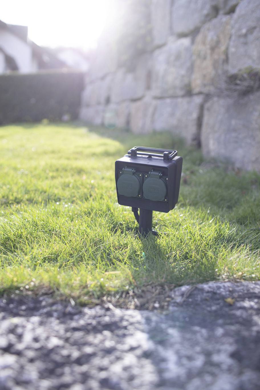 A black device with two green sockets is standing in the grass in front of a stone wall in sunlight.