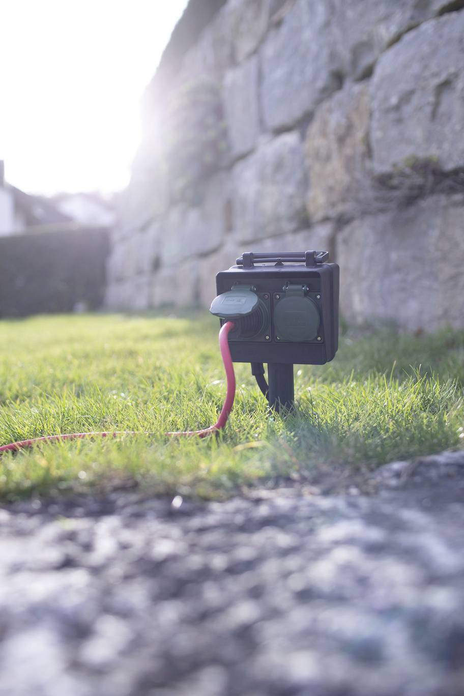 A double outdoor socket is installed on a lawn in front of a stone wall, with a red cable plugged in.