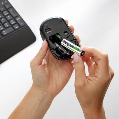 A hand is inserting a battery into an open wireless computer mouse. A keyboard is slightly out of focus in the background.