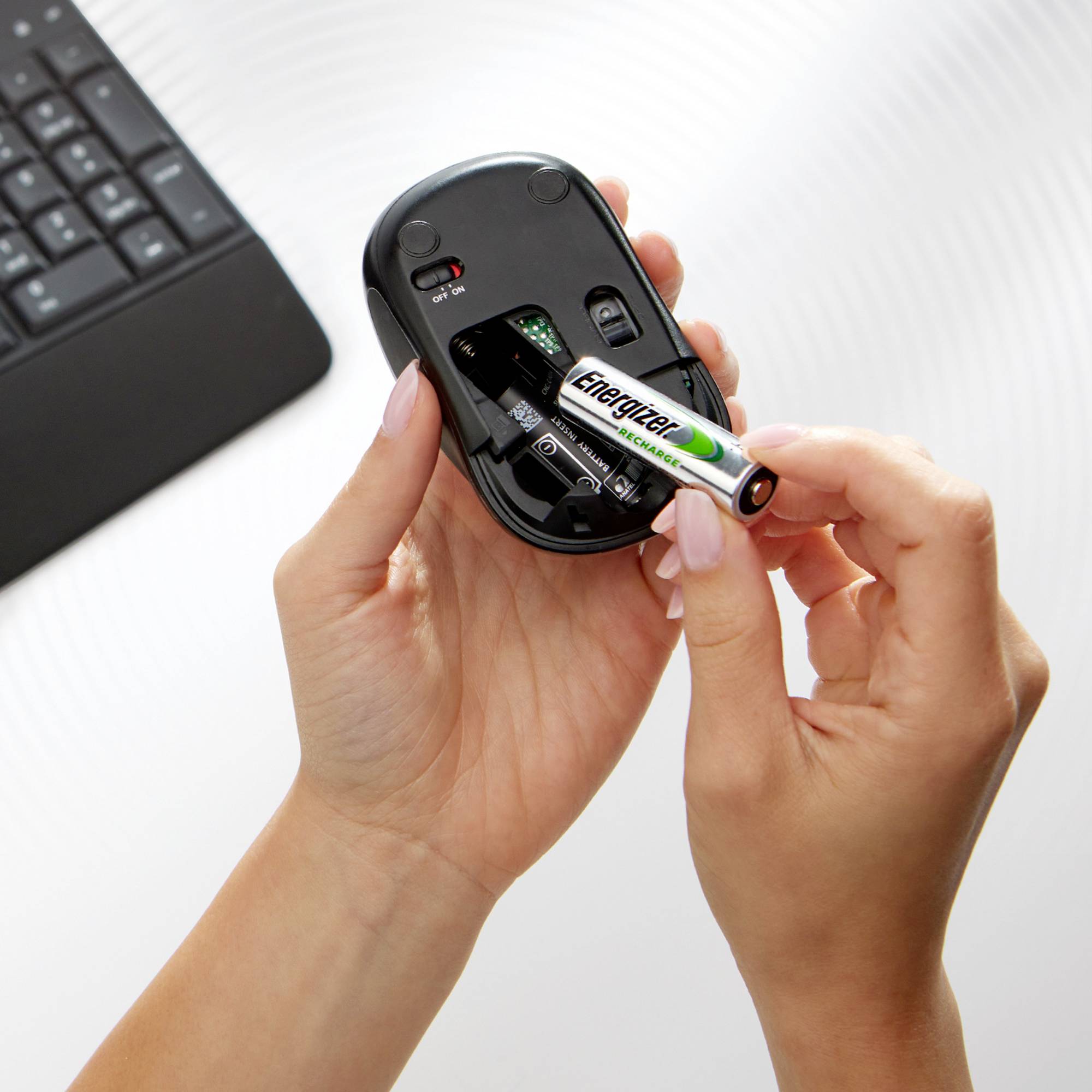 A person is holding a computer mouse and inserting a battery, next to a keyboard on a white table.