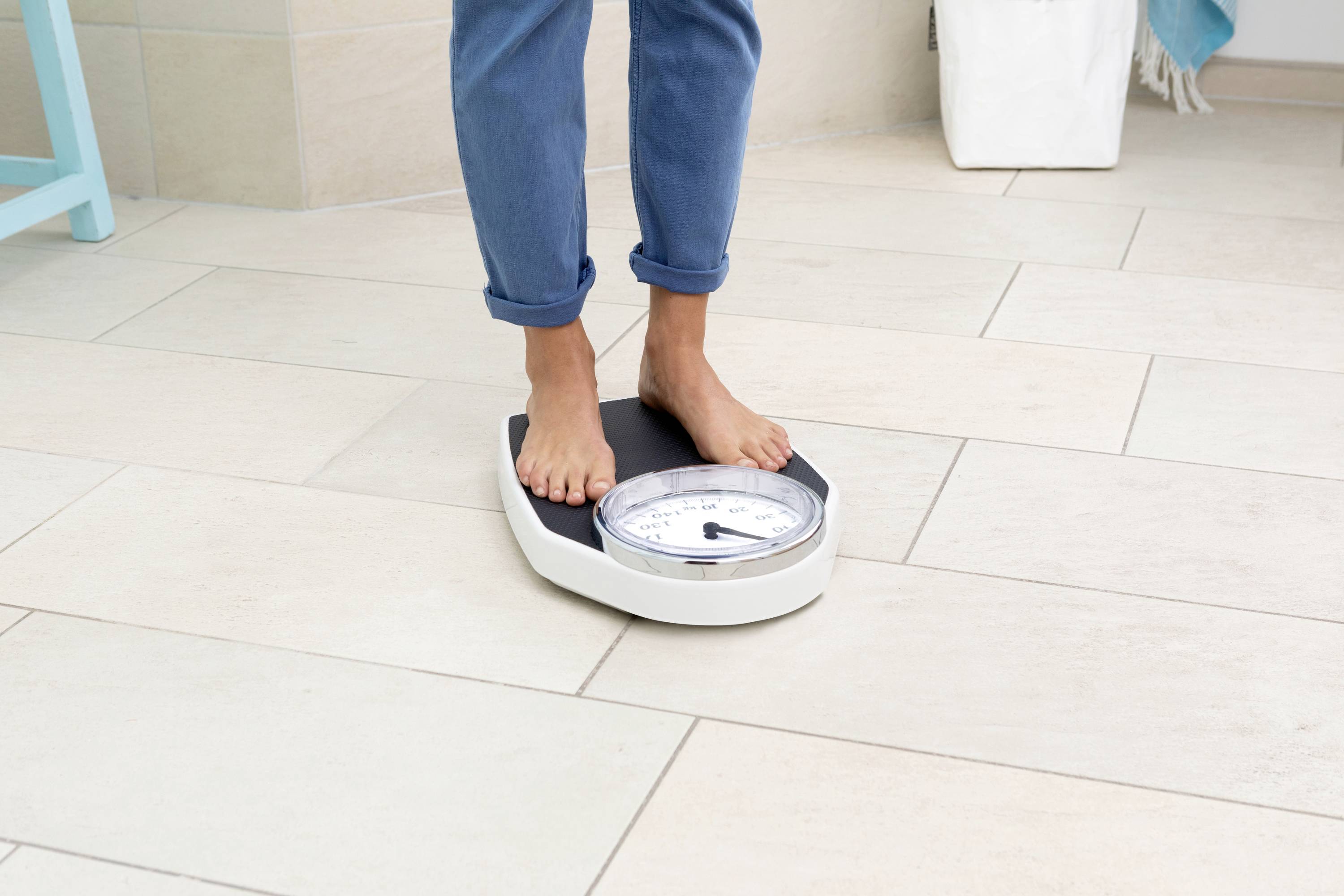 A person is standing on an analogue scale on light-coloured tiles, wearing blue jeans.