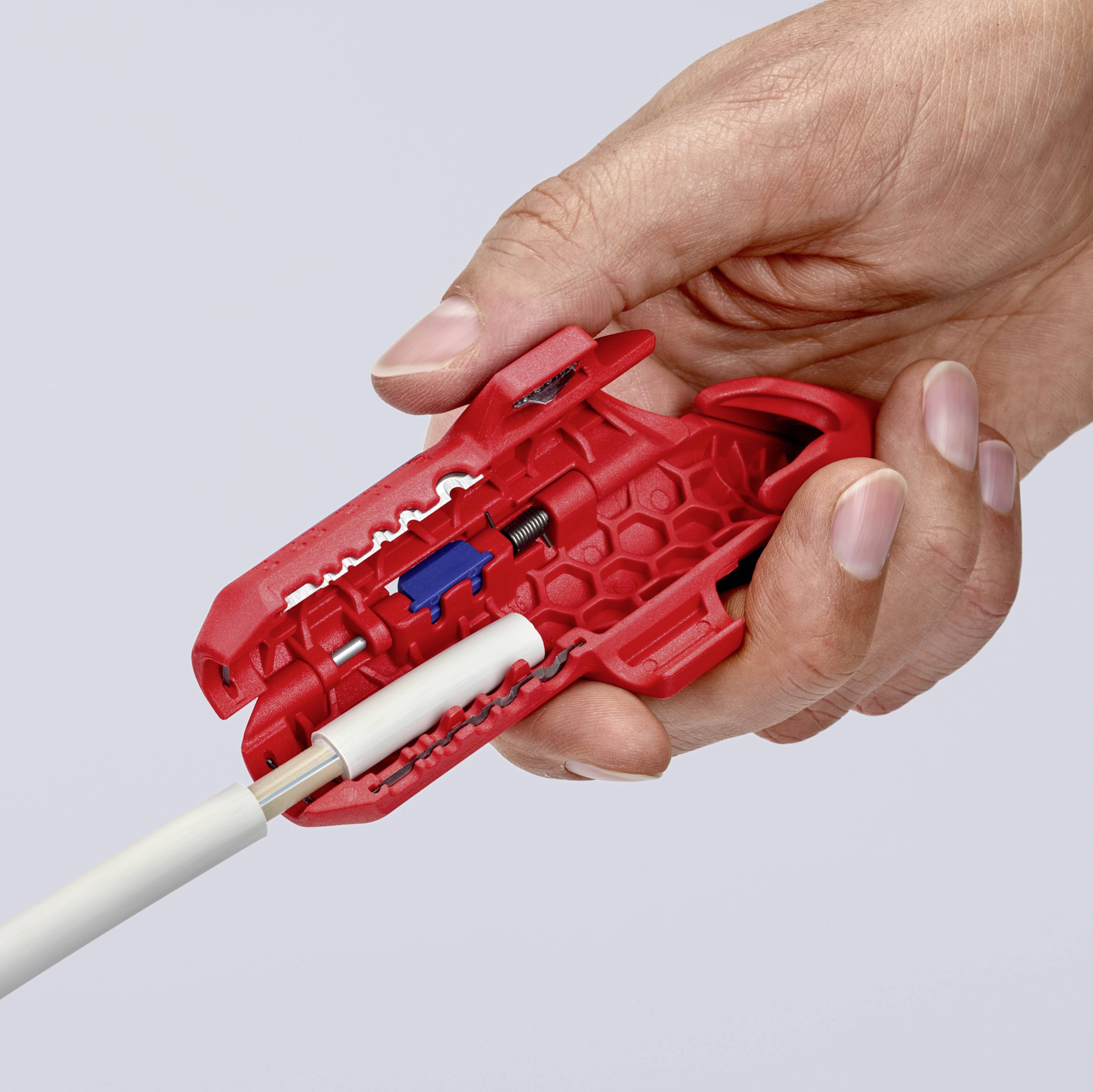 A hand holds a red tool cutting a white cable sheath. The tool reveals its internal mechanism with blue components.