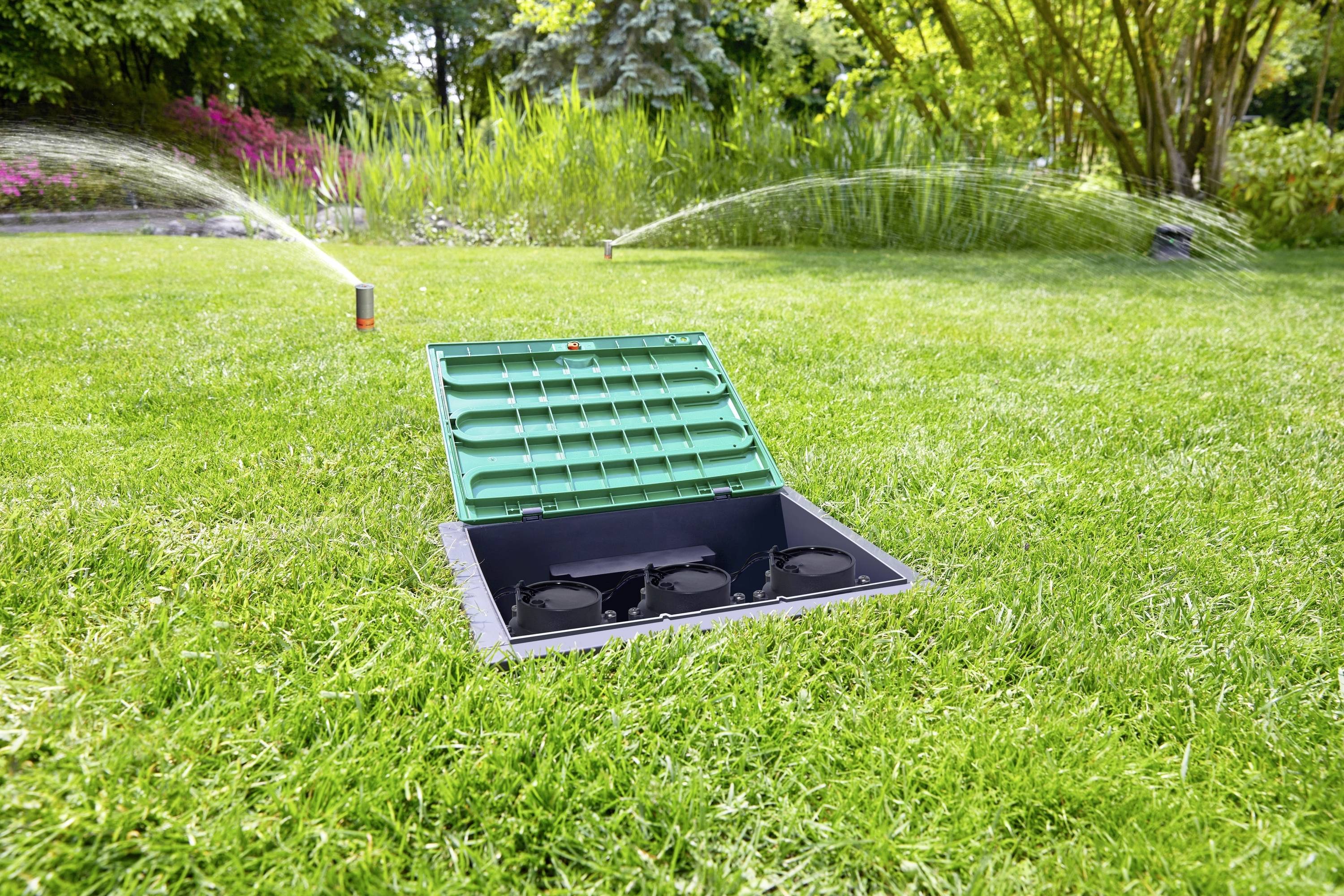 An open green valve box on a lawn shows irrigation valves. Two active lawn sprinklers can be seen in the background.