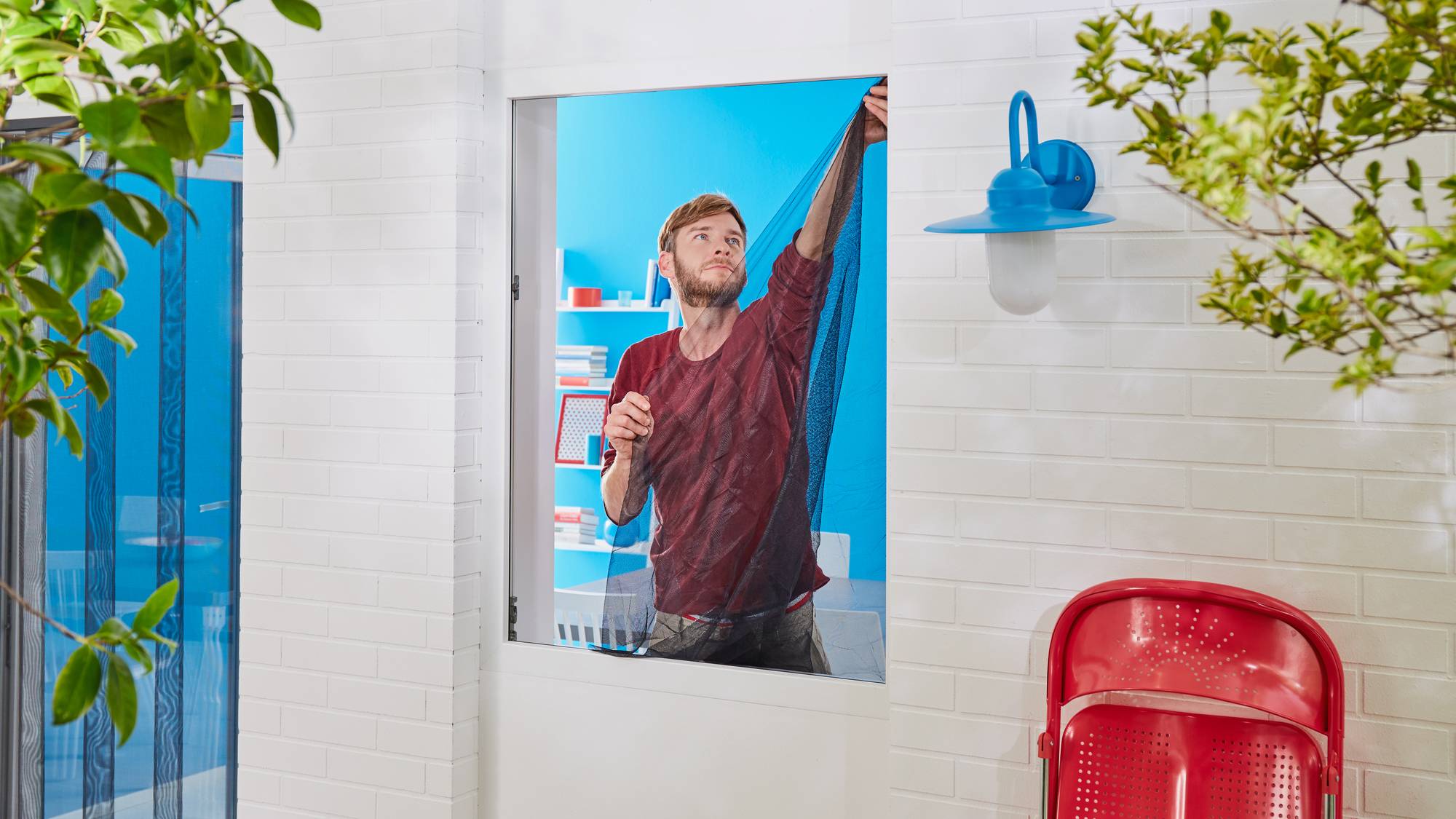 A man draws a curtain in a bright room with red chairs. Outside, a blue lamp is positioned on the white wall.