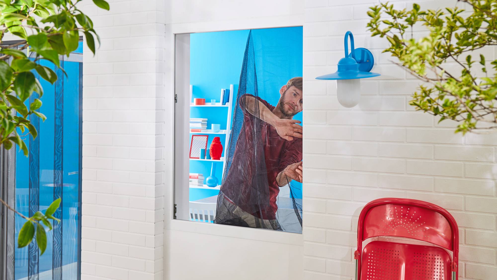 A man is securing a window screen in front of an open window. Shelves with various objects can be seen in the background.