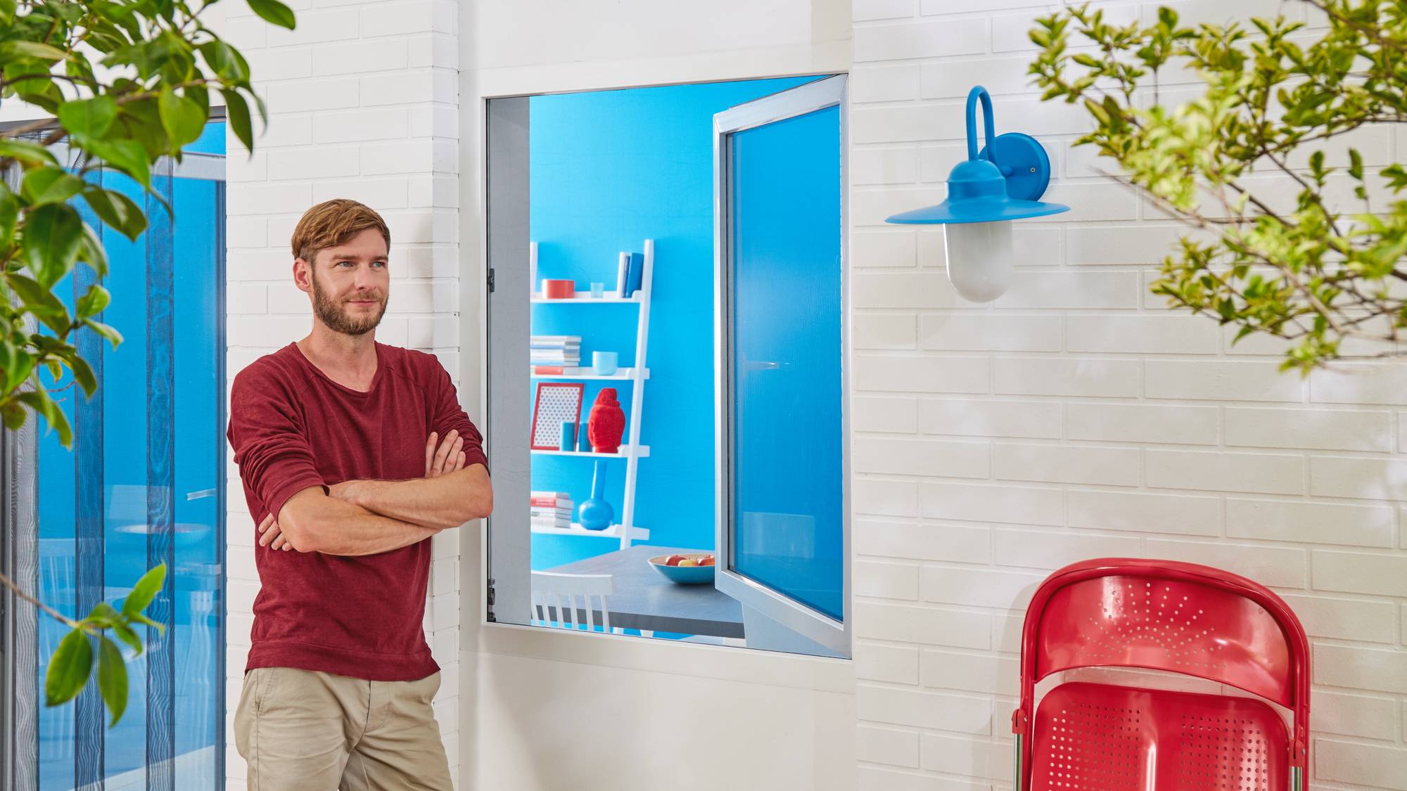 A man stands with his arms crossed in front of a white building. An open window reveals a blue room equipped with shelves.