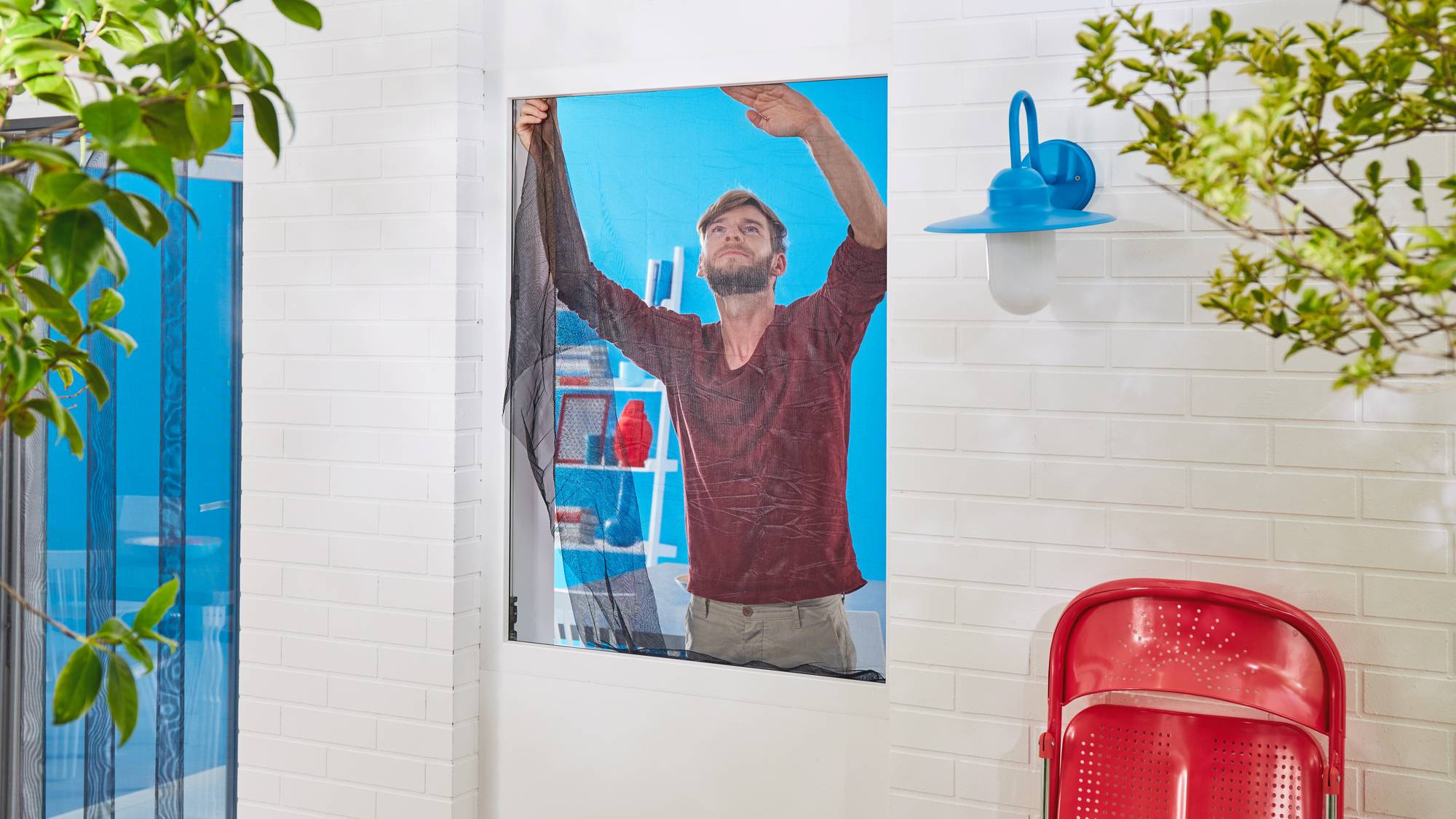 A man is fixing a mosquito net to a window. Blue walls and a red chair are visible in the background.